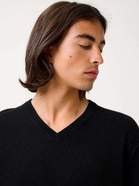 Schwarzer V-Ausschnitt-Pullover aus weichem Material, mit klassischem Design und geripptem Halsabschluss. Das Model hat schulterlanges Haar und schaut zur Seite.