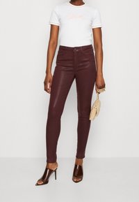 Kvinna i vit kortärmad topp, högmidjade bourgogne läderbyxor, bruna sandaler med öppen tå, håller en beige handväska med guldkedjestropp.