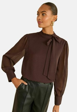 Dunkelbraune Bluse mit transparenten langen Ärmeln, hohem Kragen mit dekorativem Besatz und einer vorderen Schleifenbindung. Kombiniert mit schwarzen Lederhosen.