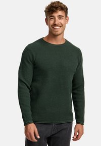 Pull en maille texturée vert avec des manches longues et un col rond, associé à un jean foncé. Possède un ourlet et des poignets côtelés.
