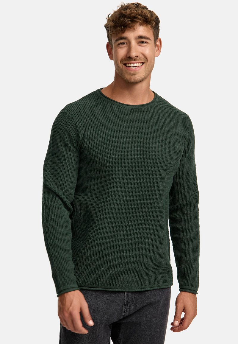 Pull en maille texturée vert avec des manches longues et un col rond, associé à un jean foncé. Possède un ourlet et des poignets côtelés.