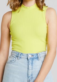 Rippenshirt ohne Ärmel in leuchtendem Limettengrün mit hohem Kragen. Kombiniert mit hellblauen, hochtaillierten Jeansshorts.