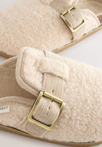 Beżowe kapcie z polaru o miękkiej fakturze, z beżowym paskiem materiałowym i złotą metalową klamrą. Lekka i swobodna konstrukcja.