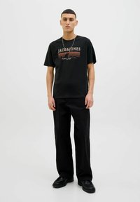 Melna T-kreka ar "JACK & JONES" logotipu oranžā un baltā krāsā, apvienota ar melnām biksēm un melniem zābakiem, ar brīvu piegriezumu un īsām piedurknēm.
