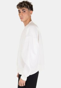 Felpa bianca oversize con scollatura rotonda, spalle scese e polsini a coste. Abbinata a pantaloni scuri, su uno sfondo neutro.