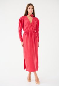 Robe rouge à manches longues en tissu doux, avec un décolleté en V profond et une taille froncée, présentant une fente sur le côté et un accent de fleur en tissu au niveau du col.