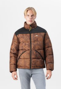 Chaqueta acolchada marrón con patrón floral, acentos en negro en los hombros, cuello alto y cierre de cremallera. Presenta bolsillos laterales y una textura suave.