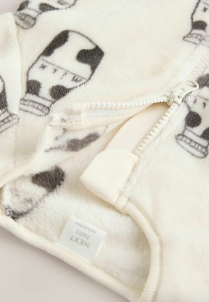 Romper per baby in morbido fleece color crema, caratterizzato da una fantasia a stampa mucca, chiusura con zip frontale e etichetta con scritto "NEXT baby."