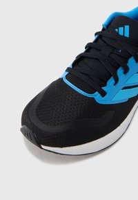Μαύρο αθλητικό παπούτσι με πλέγμα στην επάνω επιφάνεια, μπλε λεπτομέρειες, επίπεδα κορδόνια και υφή στη σόλα. Διαθέτει λαστιχένια φτέρνα και λογότυπο στη πλαϊνή πλευρά.