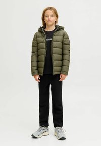 Giacca a vento verde con un design trapuntato e cappuccio, indossata sopra una maglietta nera, abbinata a pantaloni della tuta neri e sneaker grigie.