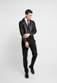 Giacca da biker in pelle nera con zip argentate, indossata sopra una camicia bianca e una cravatta nera. I pantaloni neri attillati e le scarpe eleganti nere completano il look.