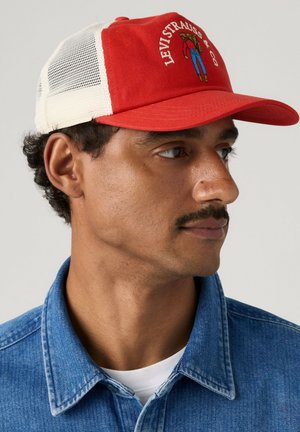 Mann trägt eine rote und weiße Levi's Trucker-Cap und eine blaue Jeansjacke, schaut nach rechts auf einem schlichten hellen Hintergrund.