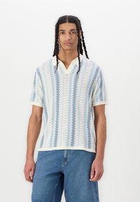 Polo majica s kratkimi rokavi v modri in beli barvi, ki ima vzorec v obliki cik-cak, V-izrez in rebraste robove. Oblečena čez jeans kratke hlače.