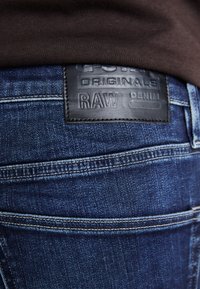 Blugi din denim albastru închis, cu o etichetă din piele neagră pe care scrie "ORIGINALS RAW DENIM", coasată aproape de talie, asortați cu o cămașă maro.