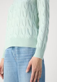Pull en maille torsadée vert clair porté avec un jean bleu clair, montrant des poignets et un ourlet côtelés, et une coupe décontractée au niveau du torse et des bras.