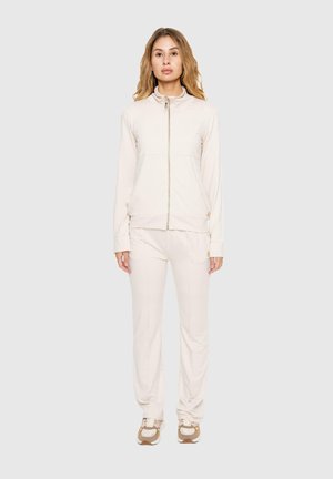 Une femme présente un sweat à capuche crème zippé, un pantalon de jogging assorti et des baskets sur un fond uni gris clair.