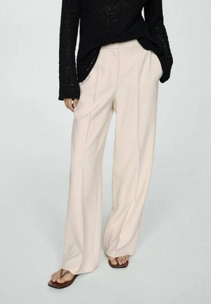 Pull en maille noir associé à un pantalon beige ample et des tongs marron, mains dans les poches, debout sur un fond clair uni.