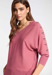 Roze gebreide trui met een V-hals en driekwartmouwen. Bevat decoratieve accenten op de schouders in verschillende kleuren. Zachte textuur.