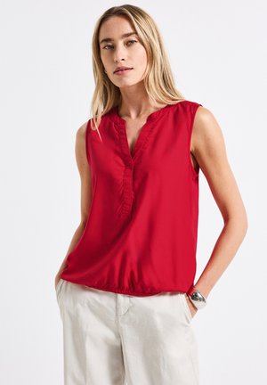 Blouse sans manches rouge en tissu lisse avec un décolleté en V, dotée de détails froncés et d'un ourlet élastiqué. Associée à un pantalon de couleur claire.