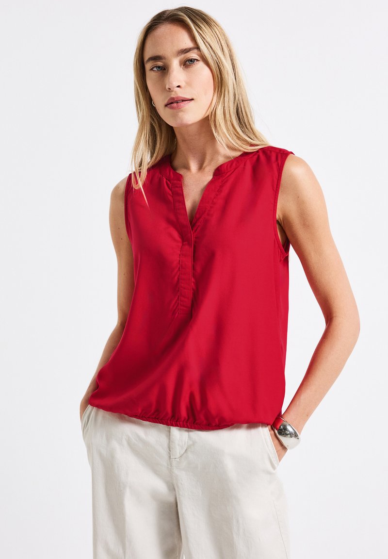Blouse sans manches rouge en tissu lisse avec un décolleté en V, dotée de détails froncés et d'un ourlet élastiqué. Associée à un pantalon de couleur claire.
