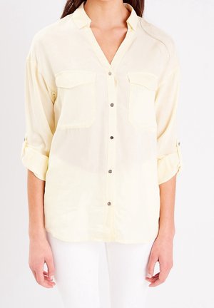 Femme portant une chemise boutonnée jaune clair à manches retroussées avec deux poches poitrine, associée à un pantalon blanc.