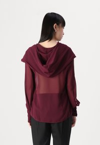 Felpa bordeaux con schiena trasparente e maniche lunghe; presenta un cappuccio drappeggiato e polsini con risvolto. Il tessuto morbido e fluente aggiunge texture.