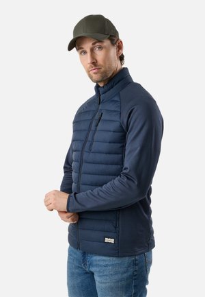 Mand iført olivengrøn kasket, marineblå quiltet jakke med lynlås og blå jeans, stående med hænderne foldet foran en hvid baggrund.