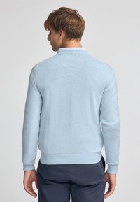 Ljusblå tröja med rund halsringning, långa ärmar, ribbade muddar och fåll. Stickad textur med slät yta. Bakvy.