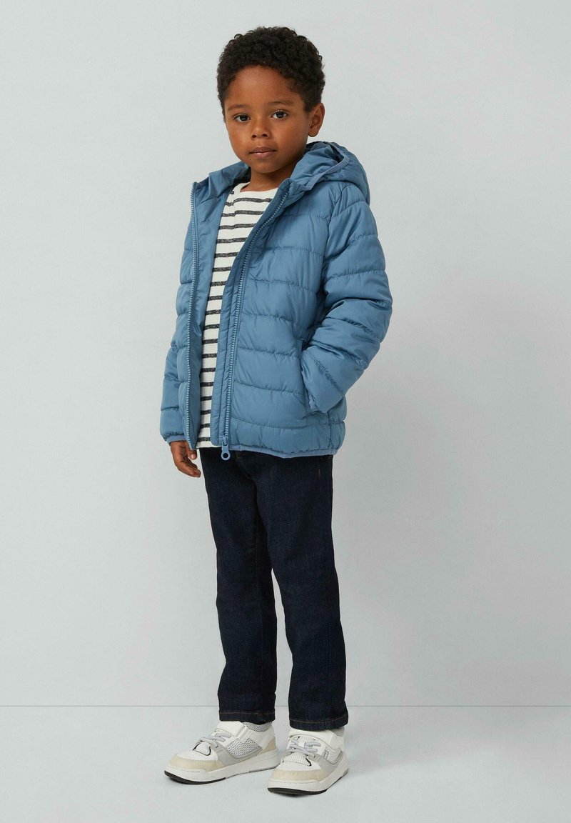 Helle blaue Steppjacke mit Kapuze, mit Reißverschluss vorne, getragen über einem gestreiften Shirt in Weiß und Marineblau. Dunkle Jeans und weiße Sneakers vervollständigen das Outfit.