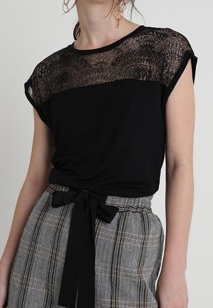 Vrouw draagt een zwart topje met kanten details op de bovenborst en schouders, gecombineerd met hooggetailleerde grijze broek met patroon, vastgemaakt met een zwarte riem.