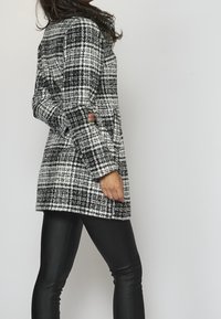 Černobílý károvaný kabát s texturovanou tkaninou, dlouhými rukávy a mírně oversize střihem. Kombinován s černými užšími kalhotami.