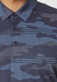 Granatowa koszulka polo z niebieskim wzorem kamuflażu, trzema guzikami i haftowanym logo na piersi.