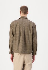 Checkered brown shirt with a relaxed fit, long sleeves, and a back pleat detail. The fabric has a textured appearance, complementing beige pants.