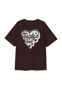 Tummanruskea puuvillapaita, jossa on valkoinen graafinen suunnittelu sydämistä ja teksti "Nostalgia Feels" selässä. Lyhyet hihnat, rento malli.