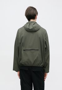 Πράσινο ελαιόχρωμο πουλόβερ-ζακέτα με κουκούλα, που διαθέτει τσέπη με φερμουάρ στο πίσω μέρος και ελαστικές μανσέτες, κατασκευασμένο από ελαφρύ, υφή υλικό.