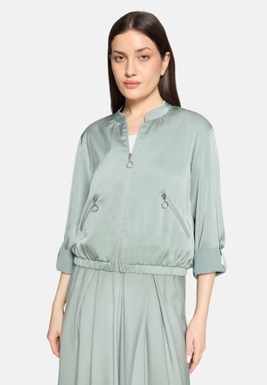 Femme portant une veste bomber en satin vert clair avec poches zippées et manches retroussées aux trois quarts, assortie à un pantalon plissé dans la même couleur.