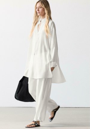 Frau mit langen blonden Haaren geht, trägt ein weißes, lockeres Hemd und Hose, schwarze Sandalen, trägt eine große schwarze Tasche vor einfachem Hintergrund.