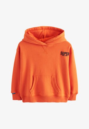 Barn med krøllet hår står med hænderne i lommerne iført en orange hættetrøje, sorte bukser og hvide sneakers mod en beige stofbaggrund.