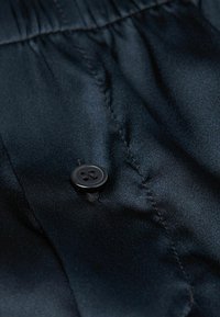 Černá saténová tkanina s jemným leskem, zdobená černým knoflíkem a vyšitými detaily. Textura je hladká s jemnými nabíráním.