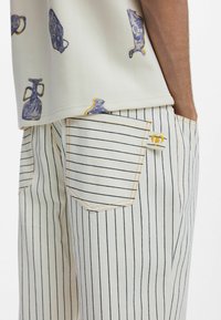 Fehér ing lila és sárga mintákkal, csíkos nadrággal párosítva, amelyen hátsó zseb és kontrasztos varrási részletek találhatók.