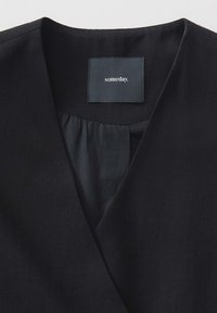 Schwarzes Wickeloberteil mit tiefem V-Ausschnitt, glatter Textur und gerafftem Detail am Hals. Das Etikett trägt die Aufschrift "irgendwann."