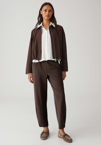 Braune Reißverschlussjacke mit Kordelzug am Saum, weißes Hemd mit Knöpfen, braune Hose und braune Slipper. Glatte Texturen und maßgeschneiderte Passform.