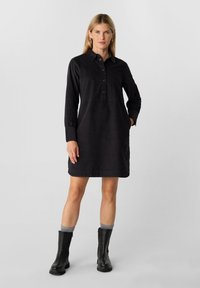 Schwarzes Hemdkleid mit langen Ärmeln, kragenloser Ausschnitt, Knopfleiste und seitlichen Taschen. Kombiniert mit schwarzen Stiefeletten und grauen Socken.