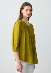 Donna con lunghi capelli castani, che indossa una blusa larga di un verde oliva strutturato con colletto ricamato e pantaloni bianchi, in piedi su uno sfondo semplice.