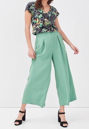 Femme portant un haut noir à manches courtes à motifs floraux, un pantalon large taille haute vert menthe et des sandales noires à talon bloc avec bride à la cheville, debout sur fond blanc.