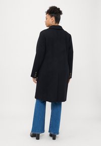 Fekete gyapjúkeverék kabát, lekerekített hajtókával, egyenes szabással és gombos mandzsettával, kék harangszár�ú farmerral és bokacsizmával viselve.