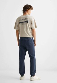 Beige bomullst-shirt med grafik på baksidan, ihop med marinblå mjukisbyxor med elastisk midja och vita sneakers med uppvikta anklar.