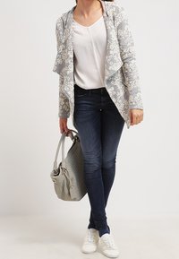 Giacca grigio chiaro con motivo floreale e davanti drappeggiato, top bianco, jeans blu scuro, sneakers bianche e borsa grigia texturizzata.
