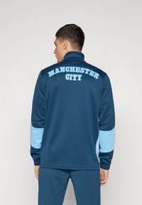 Temno modra športna jakna z svetlo modrimi poudarki, visokim ovratnikom in besedilom "MANCHESTER CITY" izstopajoče prikazano na hrbtu.