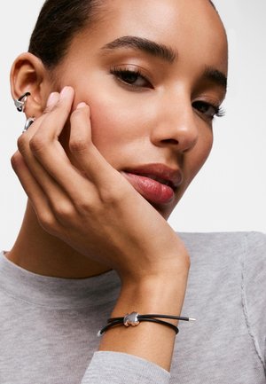 Mujer con piel suave apoyando su rostro en la mano, vestida con una camiseta gris de manga larga, pendientes de plata tipo puño y una pulsera negra con un charm plateado.